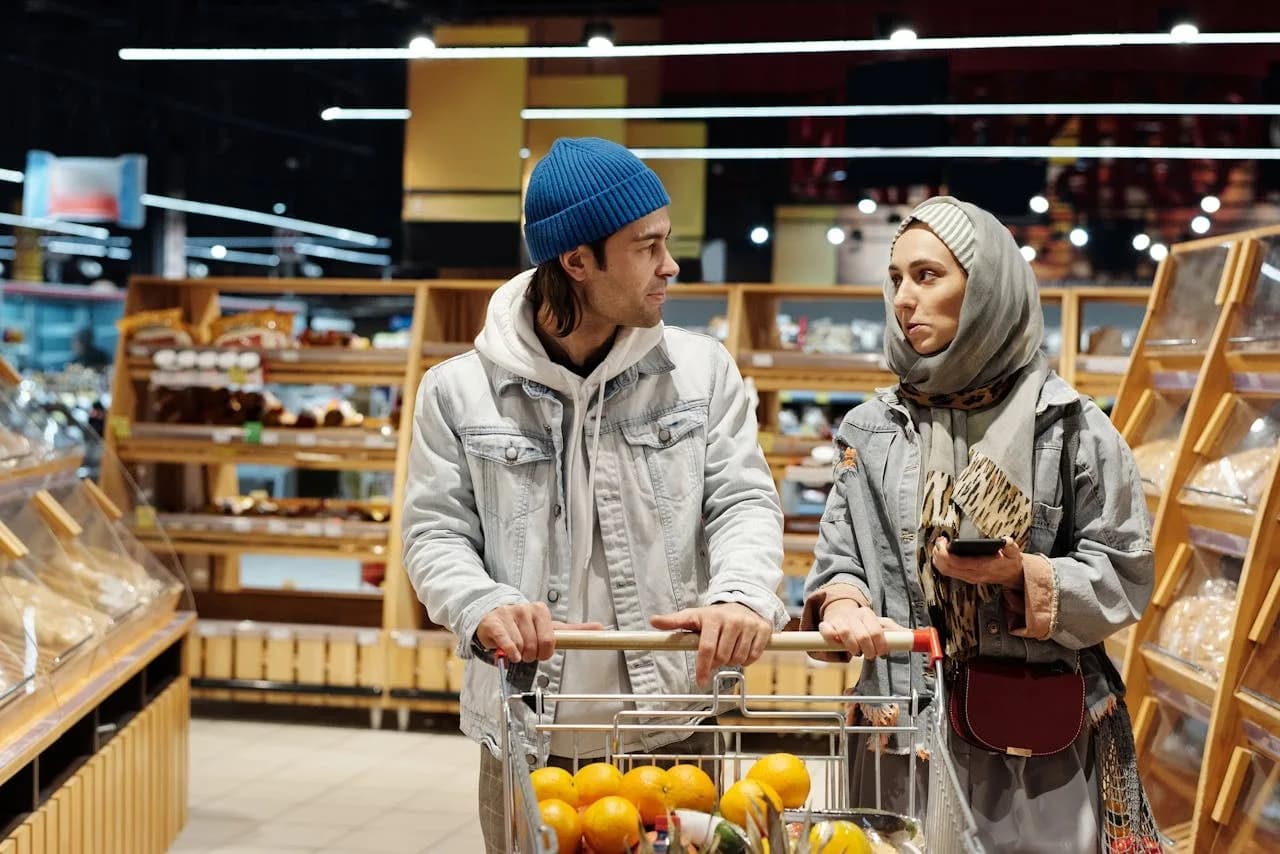 A person stands in a grocery store, holding their phone with an organized shopping list app open, systematically checking off items as they shop for planned meals. This methodical approach helps them save money and reduce stress while grocery shopping for the week.