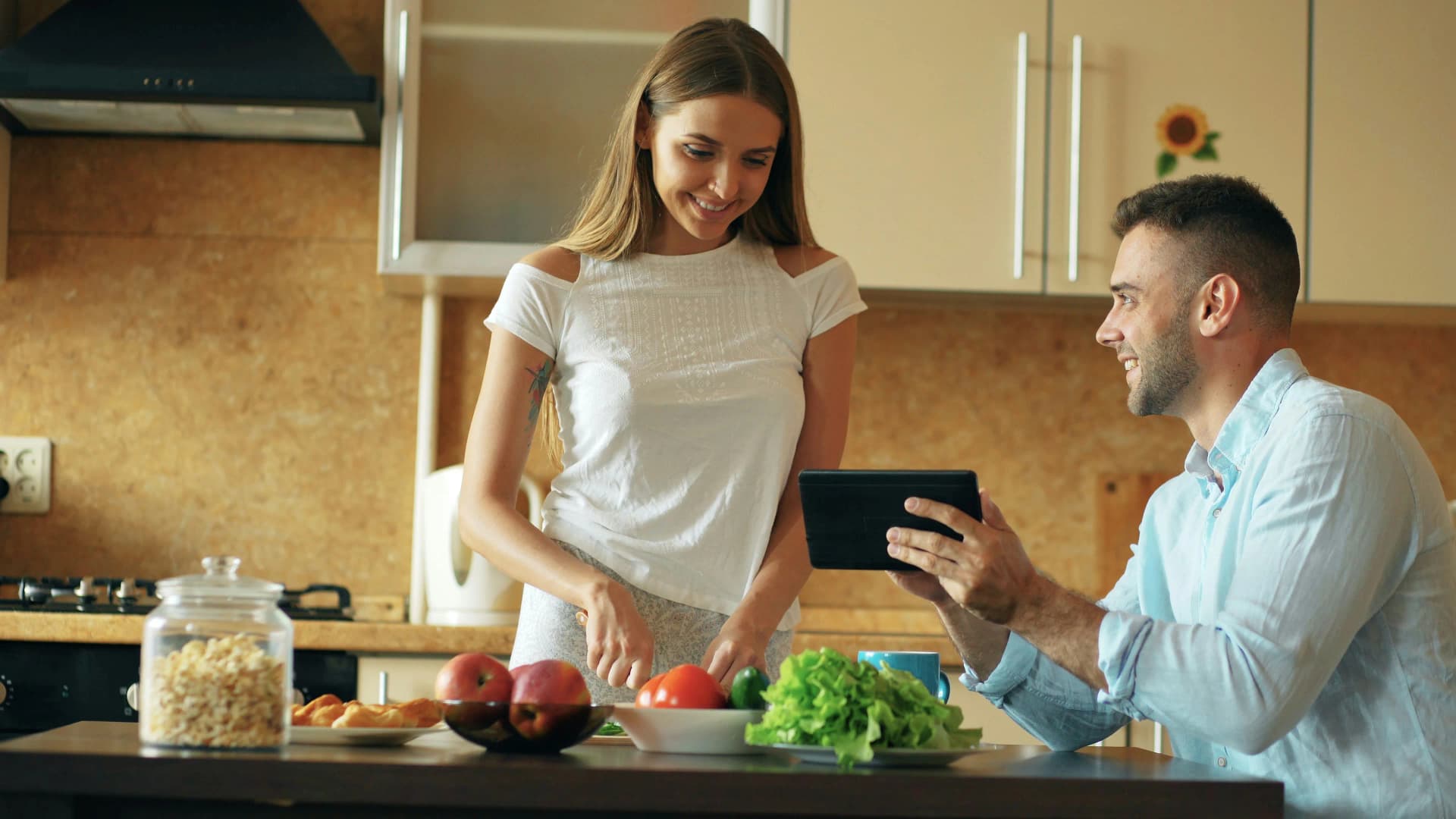 A cheerful couple sitting at a kitchen table, using Kitchendary on a tablet while groceries are spread around.